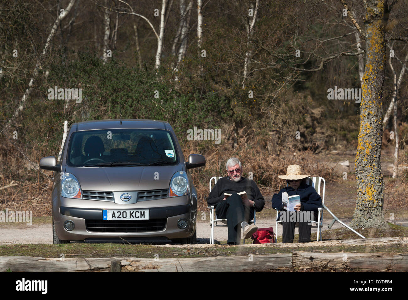 Two middle aged people in picnic chairs alongside car reading in sunshine. Stock Photo