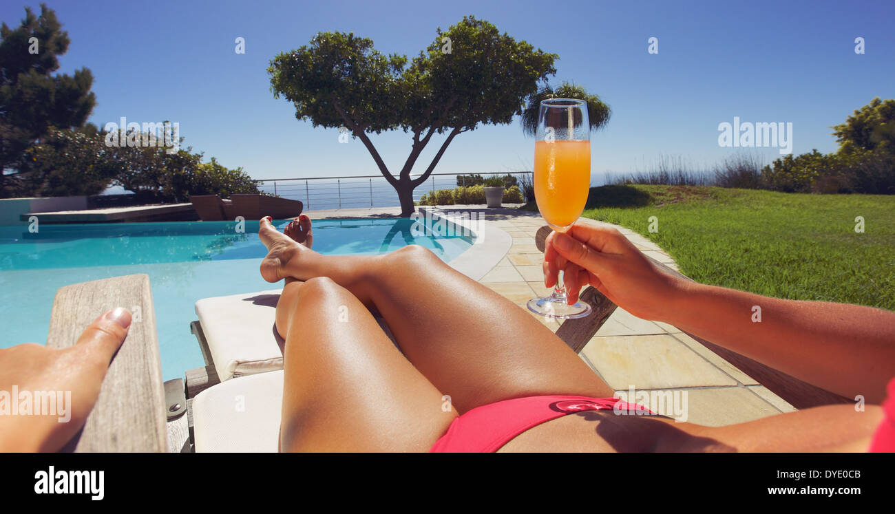 Young lady in bikini holding orange juice glass while sitting on a lounge chair along the poolside on a sunny day. Panorama view Stock Photo