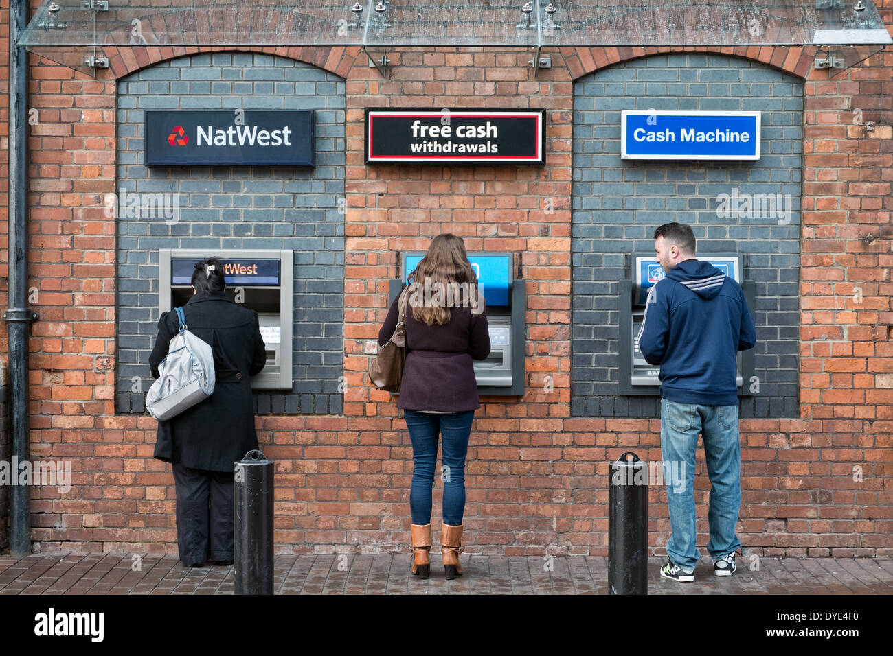 Three people at adjacent cash machines, one man watching the others. Stock Photo