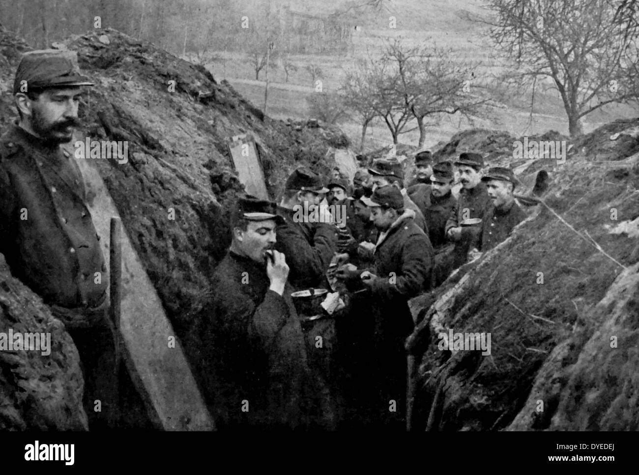 French soldiers are seen eating whilst squatting in the trenches. A soldier is seen standing guard. Dated 1914 Stock Photo