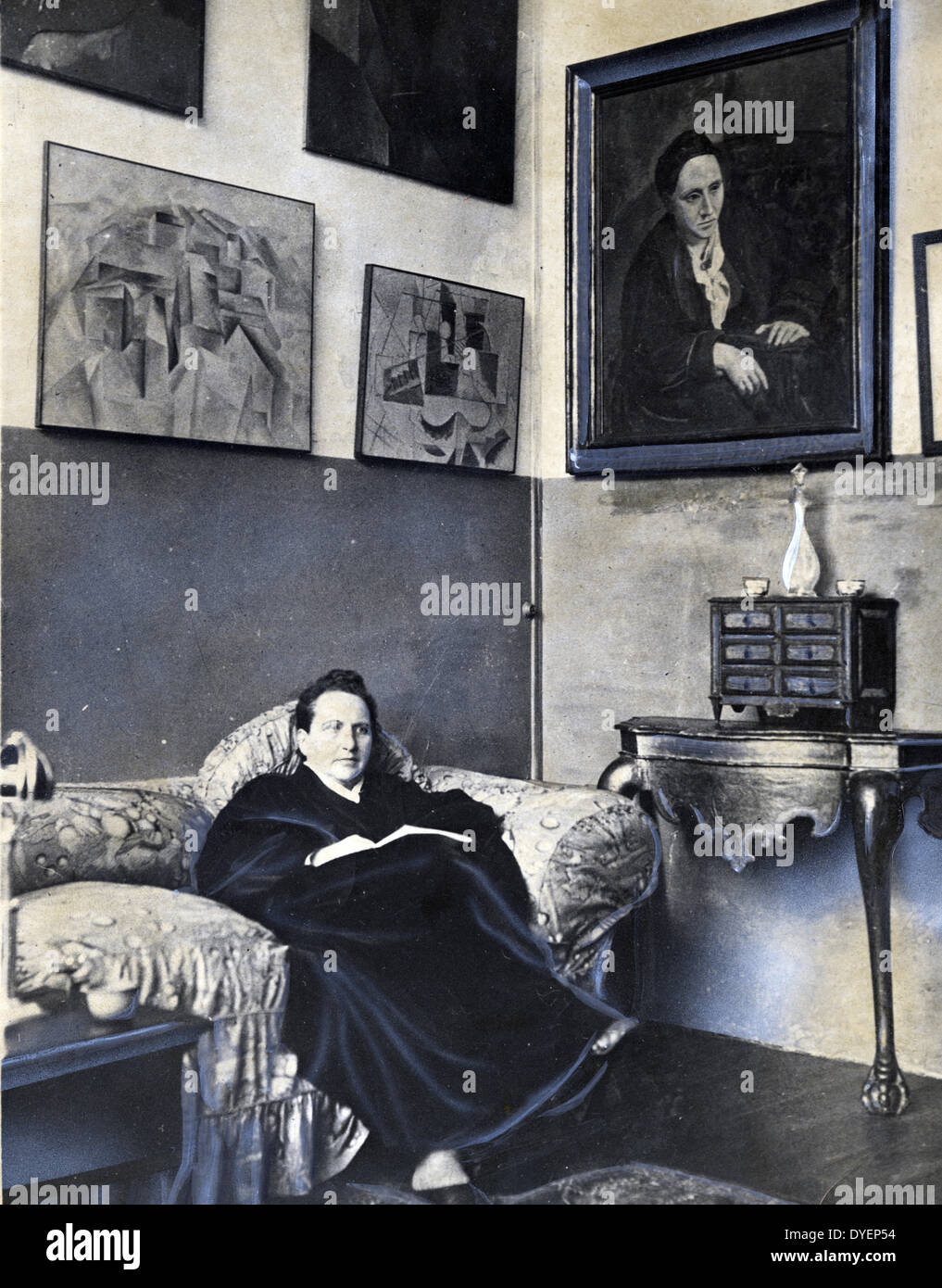 Gertrude Stein sitting on a sofa in her Paris studio, with a portrait of her by Pablo Picasso, and other modern art paintings hanging on the wall behind her 1930 Stock Photo