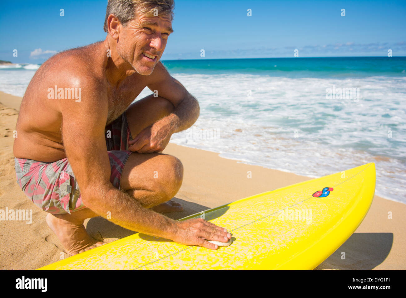 Middle-aged surfer waxing up his surfboard before heading into the surf, Hawaii Stock Photo