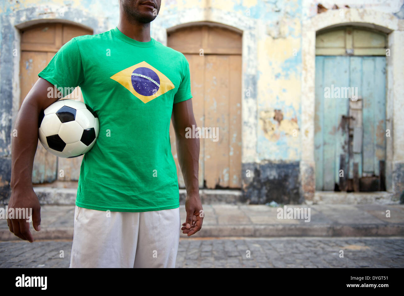 Brazilian football player stands holding soccer ball on an old rustic village street Stock Photo