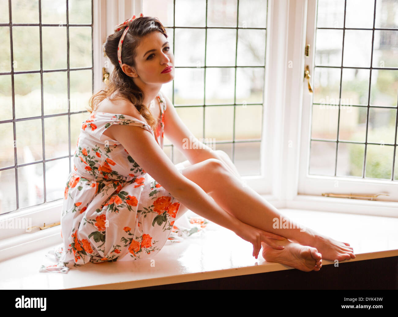 Young woman on a window sill in and old house, lit by natural light, while in a Fifties style summer dress. Stock Photo
