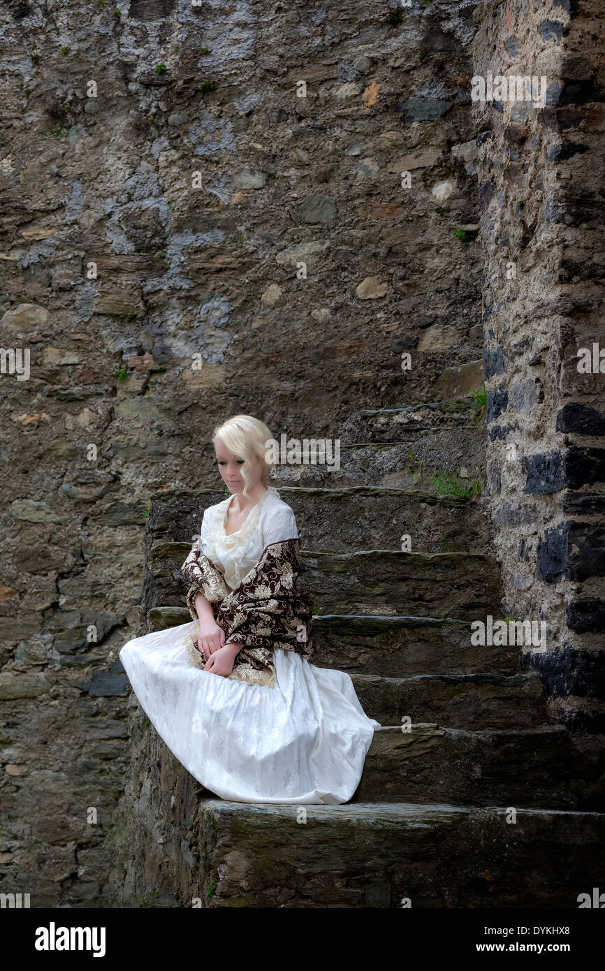 a lady in a white period dress is sitting on old stone steps Stock Photo