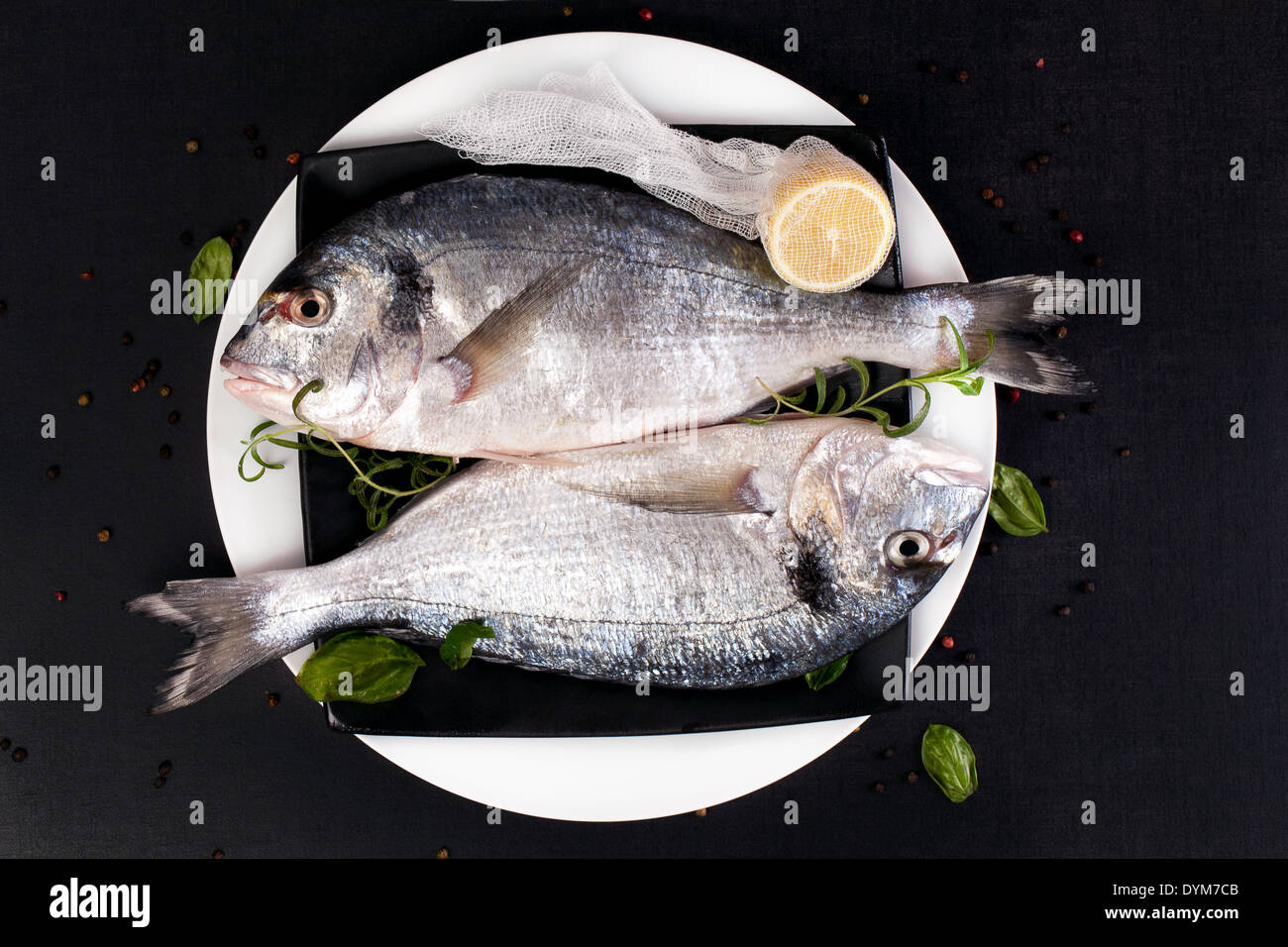 Two fresh sea bream fish on plate with fresh herbs basil, rosemary, cut lemon and colorful peppercorn, top view. Luxury fish Stock Photo