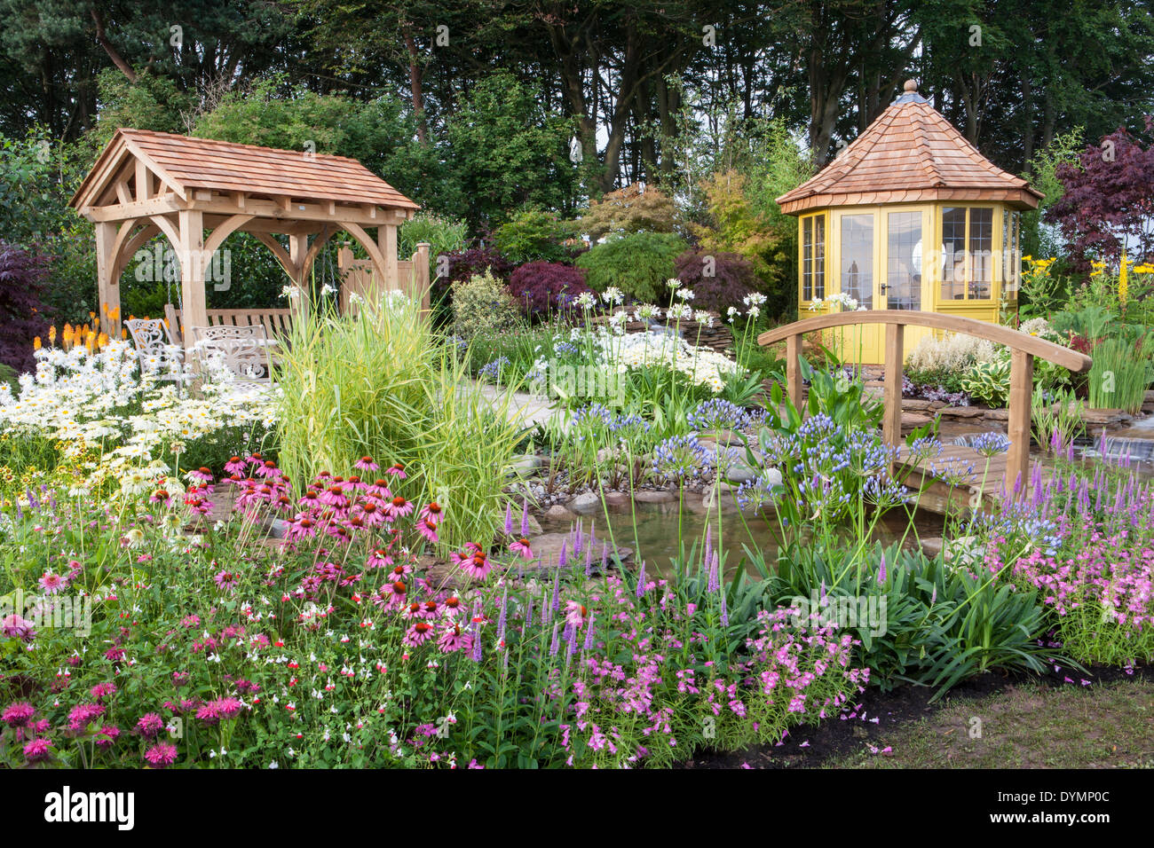 English country cottage garden arbour in summer with bridge summerhouse summer house cabin small pond water feature a mixed colourful flower border UK Stock Photo