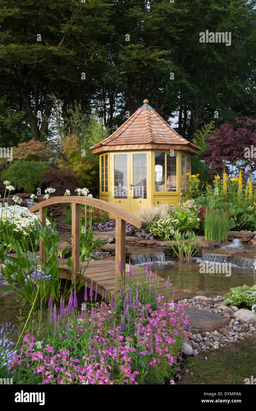 A cottage garden in summer with a bridge over to summerhouse summer house cabin small pond water feature waterfall a mixed colourful flower border UK Stock Photo