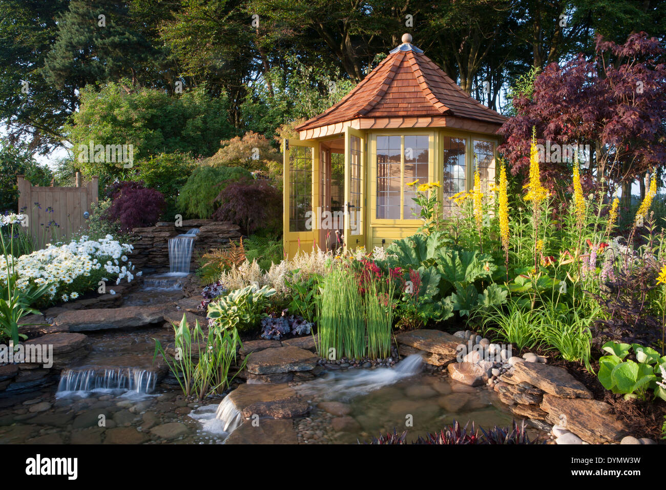A country suburban garden in summer with a summerhouse summer house cabin small pond water feature with waterfall and mixed colourful flower border UK Stock Photo