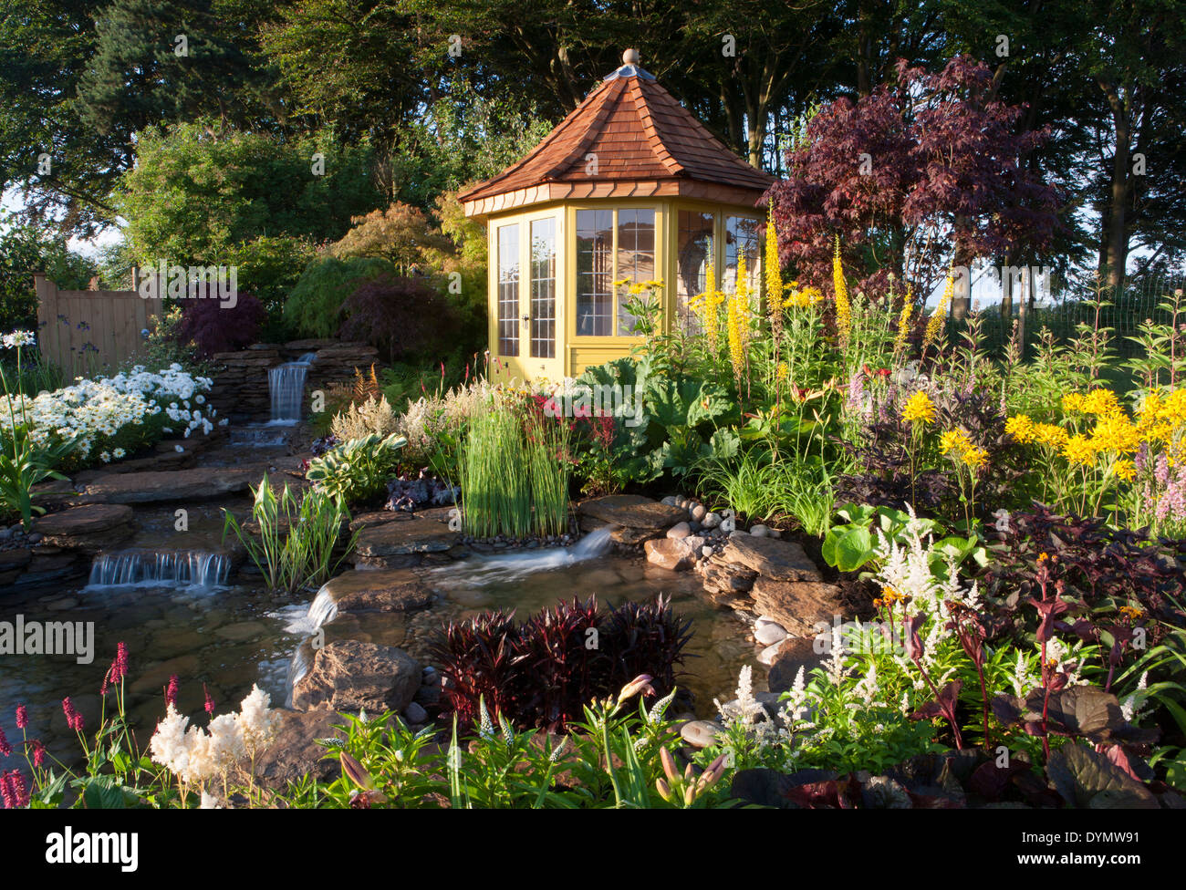 A country suburban garden in summer with a summerhouse summer house cabin small pond water feature with waterfall and mixed colourful flower border UK Stock Photo
