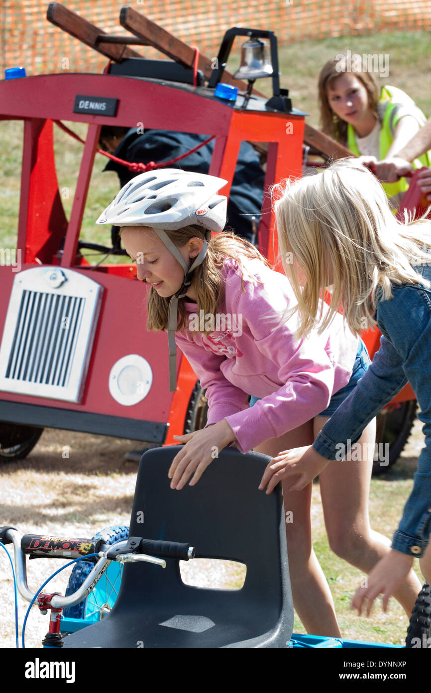 Young girl in pink top and crash helmet pushes a homemade gravity racer back up the hill, novelty fire engine in the background. Stock Photo