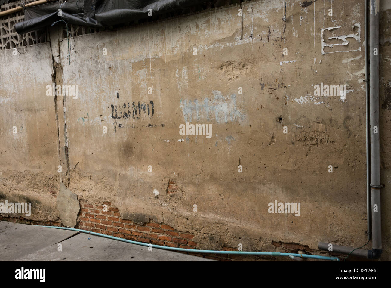 An old worn down brown wall with exposed bricks on the bottom, some Thai graffiti words written on it. Stock Photo