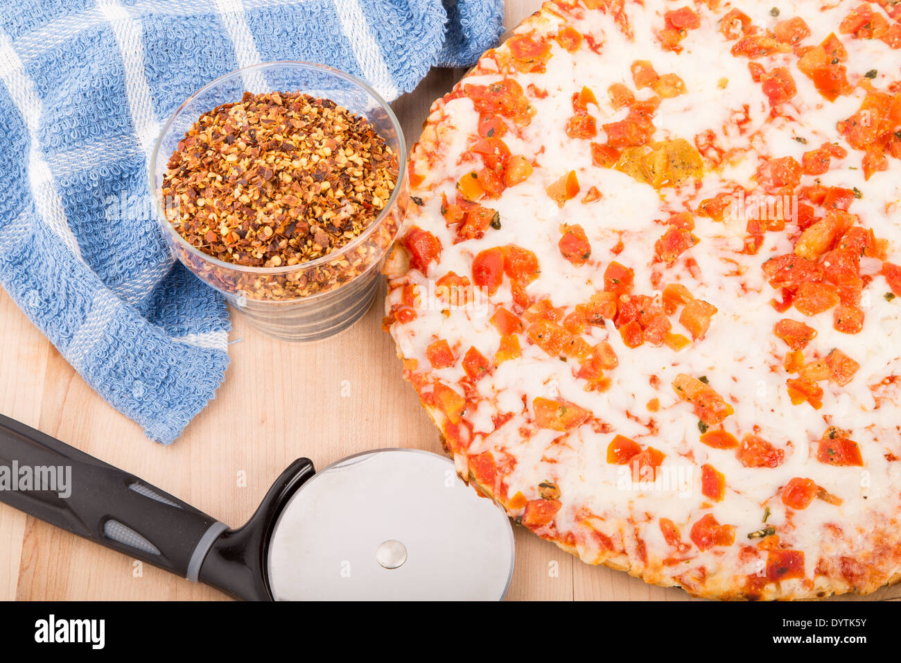 Fresh, hot tomato pizza on a wood cutting board with cutting wheel, hot pepper flakes, and a blue towel Stock Photo