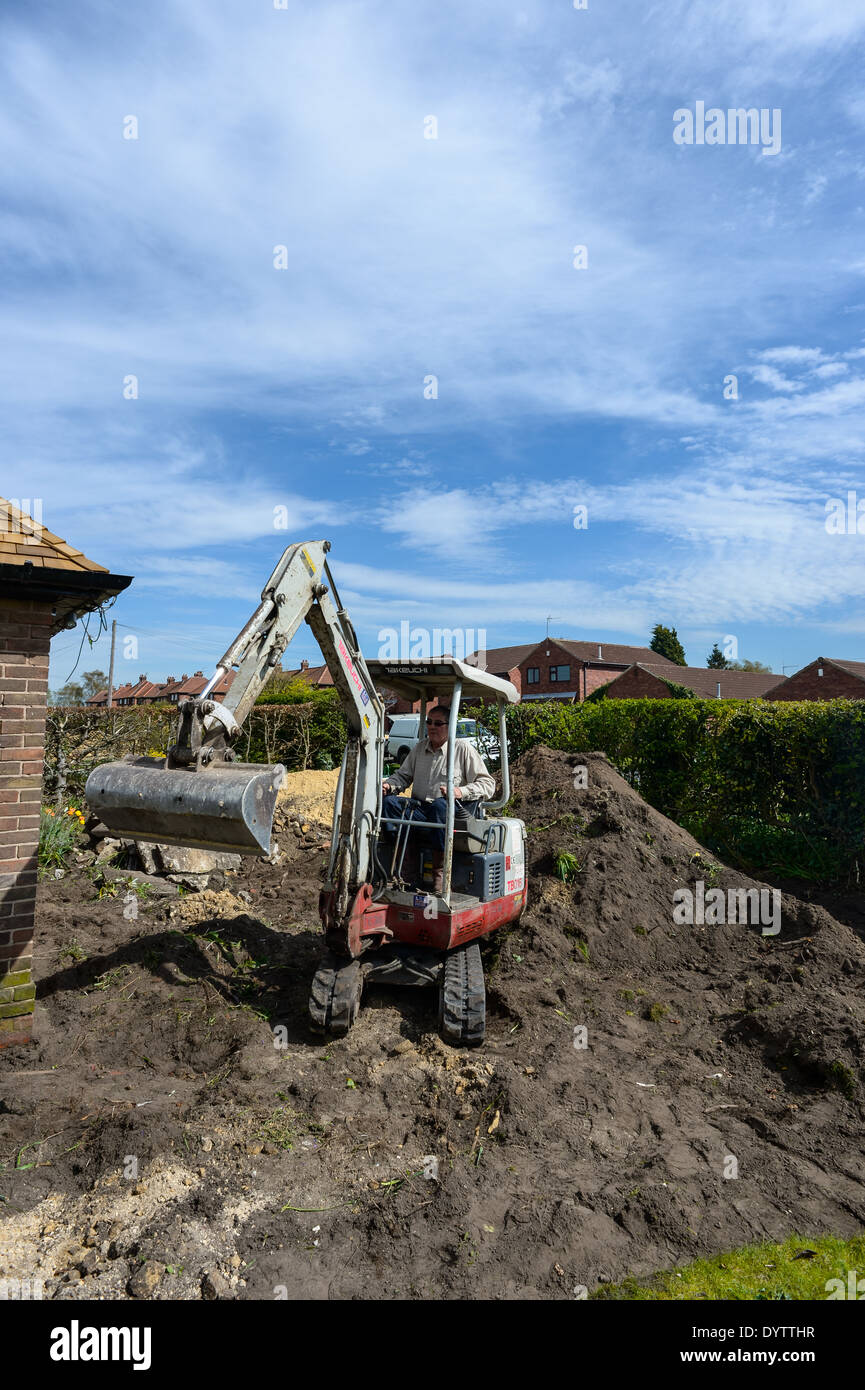 A workman digs up a garden using a micro compact excavator prior to laying a driveway on a renovation project. Wide angle view Stock Photo