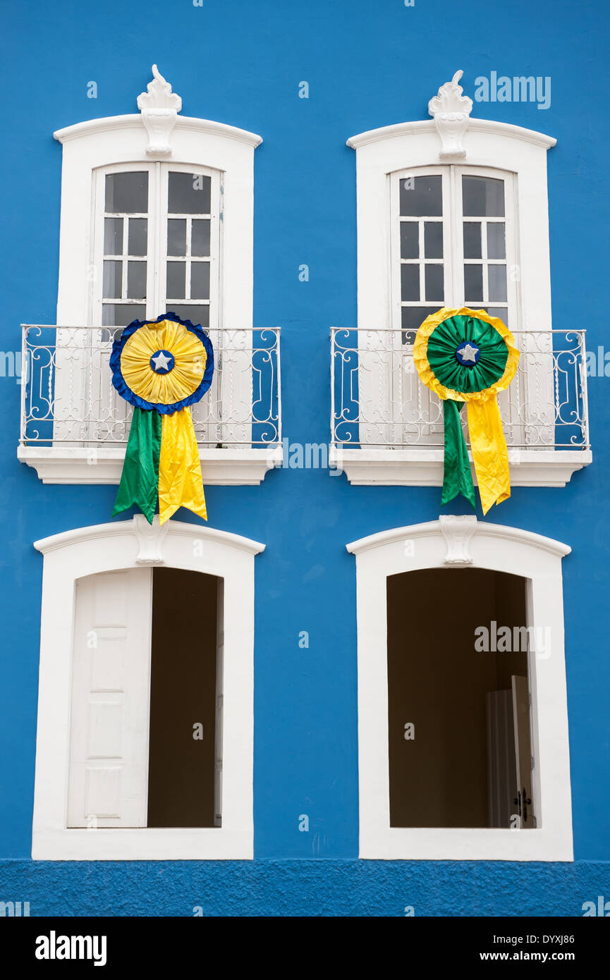 Penedo, Alagoas State, Brazil. Two rossettes in the Brazilian national colours of green, yellow and blue at the windows of a colonial building with four windows painted white in a blue wall. Stock Photo
