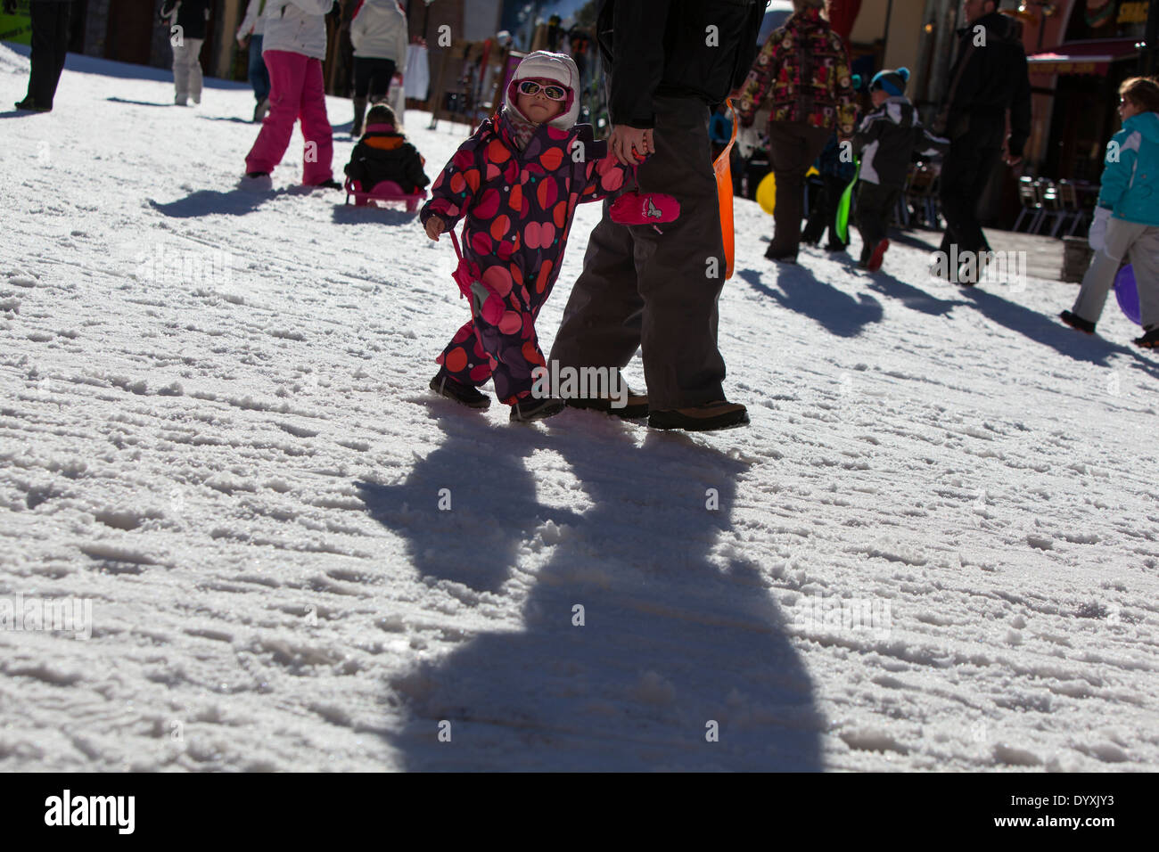 A young child dressed in a spotty all-in-one suit walks with an adult though a ski resort. Stock Photo