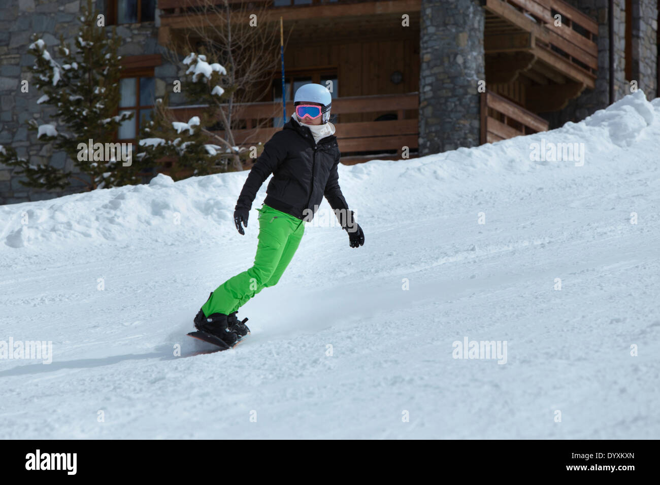 Snowboarder with bright green trousers, helmet and pink goggles, traversing the piste. Stock Photo
