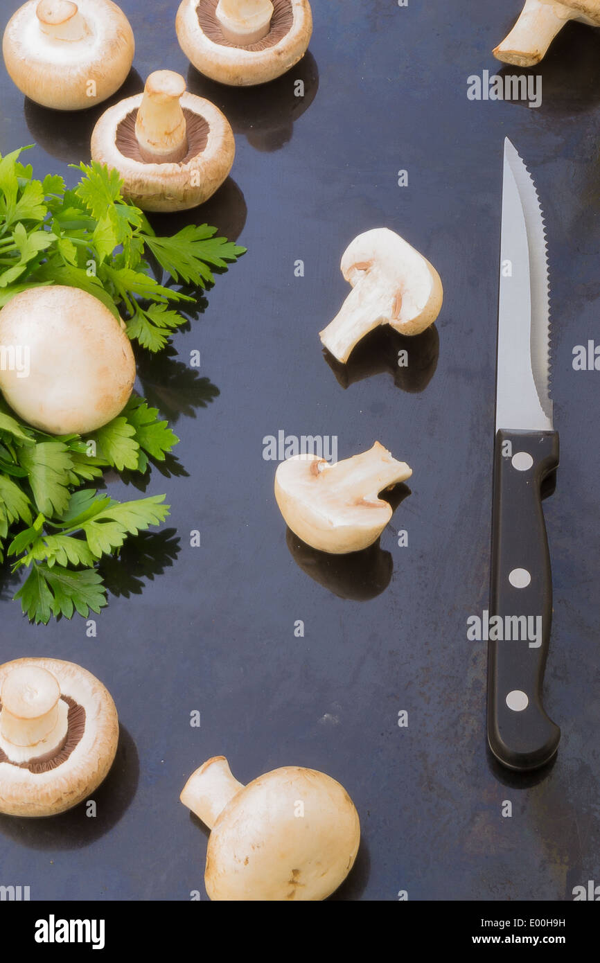 Button mushrooms, parsley and a knife spread out over the image on a blue metallic background. Stock Photo