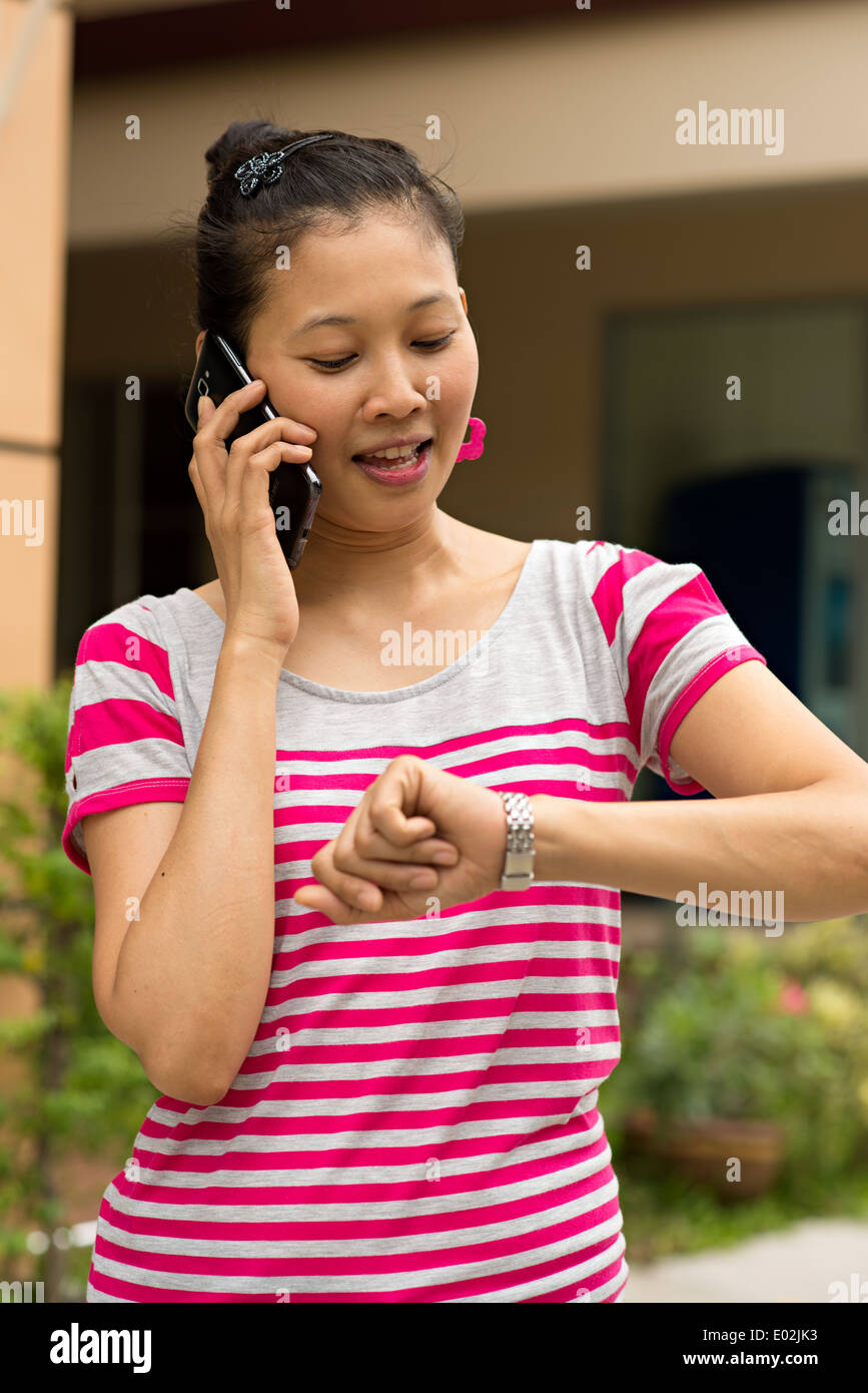 A pretty young Asian woman talking on a smart phone and smiling looking at her watch, standing outdoors. Stock Photo