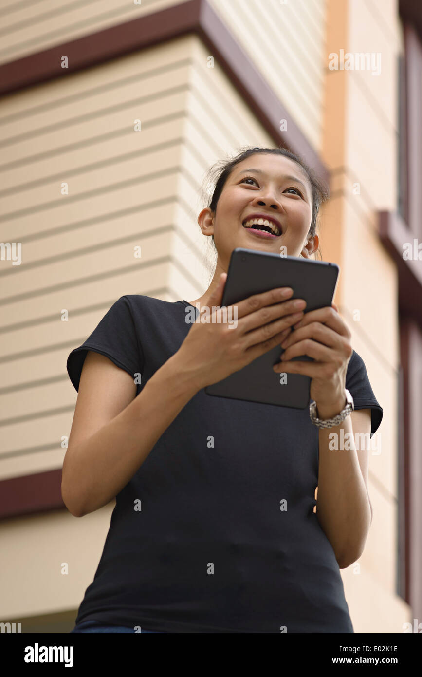 A young Asian woman holding an Ipad, tablet, smiling looking away. She wears a black T-shirt. Stock Photo