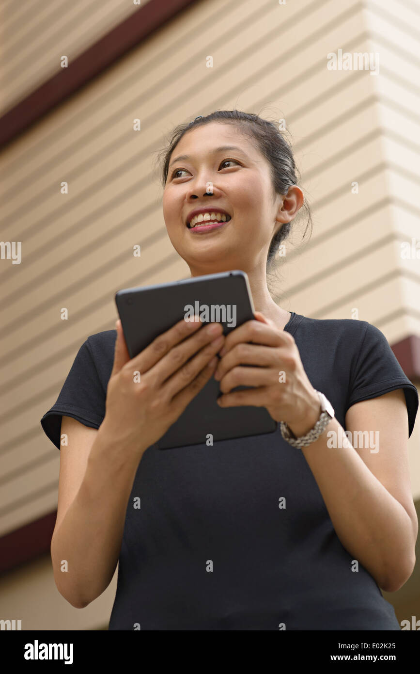 A young Asian woman holding an Ipad, tablet, smiling looking away. She wears a black T-shirt. Stock Photo