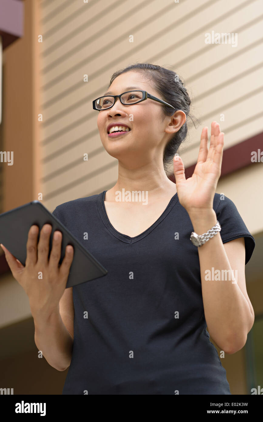 A young Asian woman wearing eyeglasses, holding an Ipad, tablet, smiling looking away waving at someone. Stock Photo