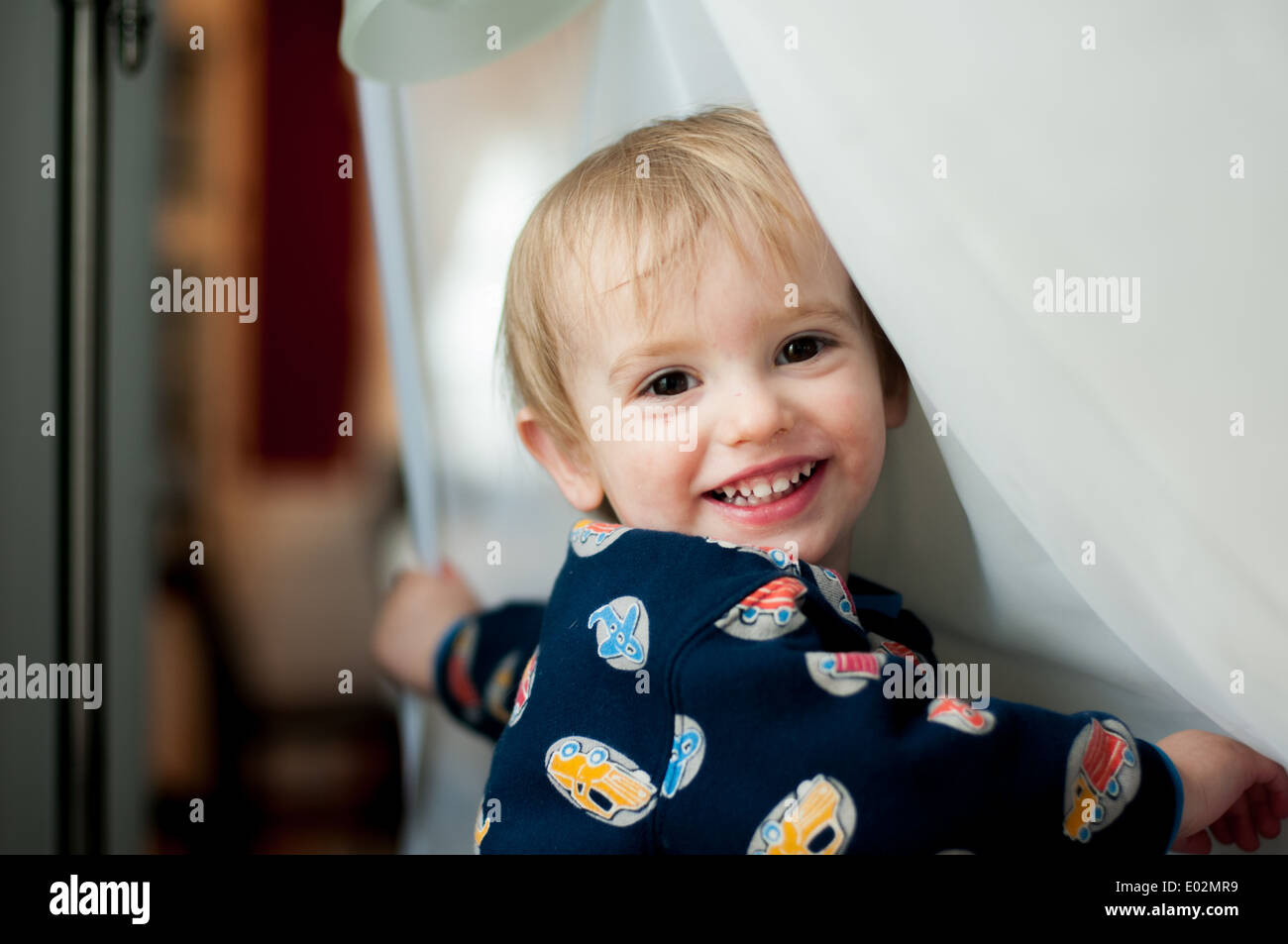 A blond toddler boy smiles while playing with a white curtain by a window. Stock Photo