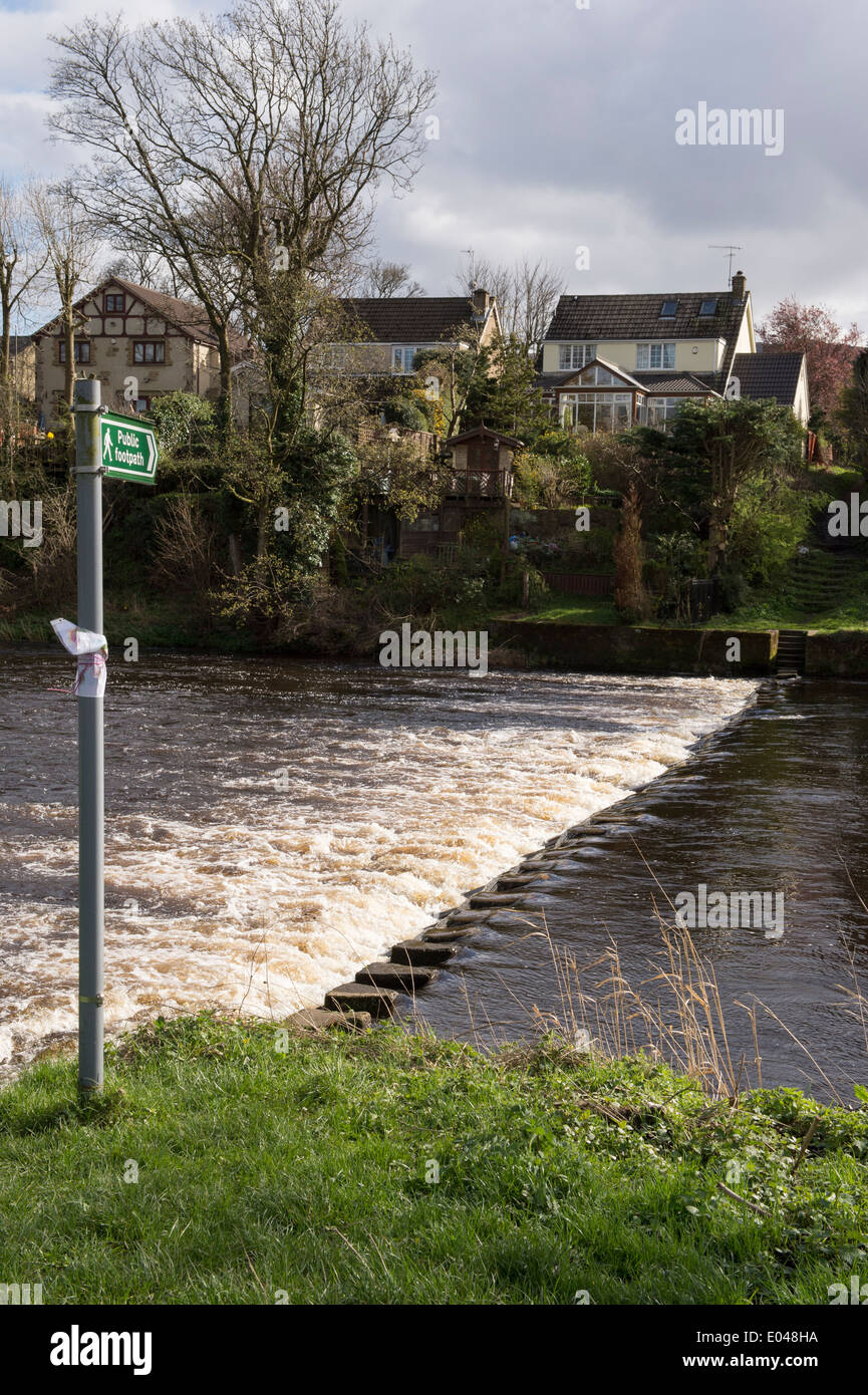 Public footpath sign pointing to partially covered stepping stones crossing flowing river - River Wharfe near Ilkley, West Yorkshire, England, UK. Stock Photo