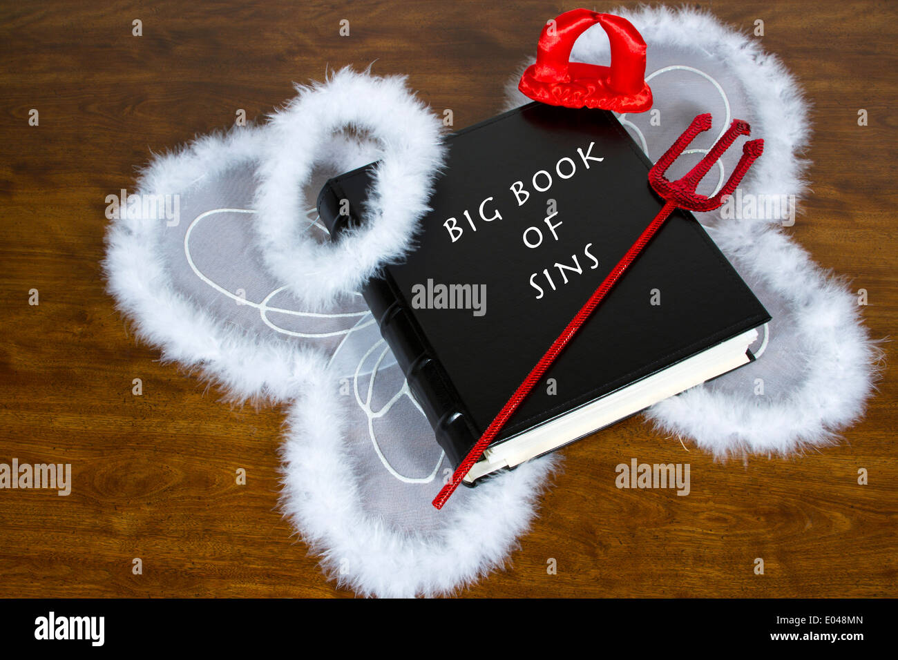 Large book labeled Big Book Of Sins on desktop with angel wings and halo, devil horns and pitch fork Stock Photo