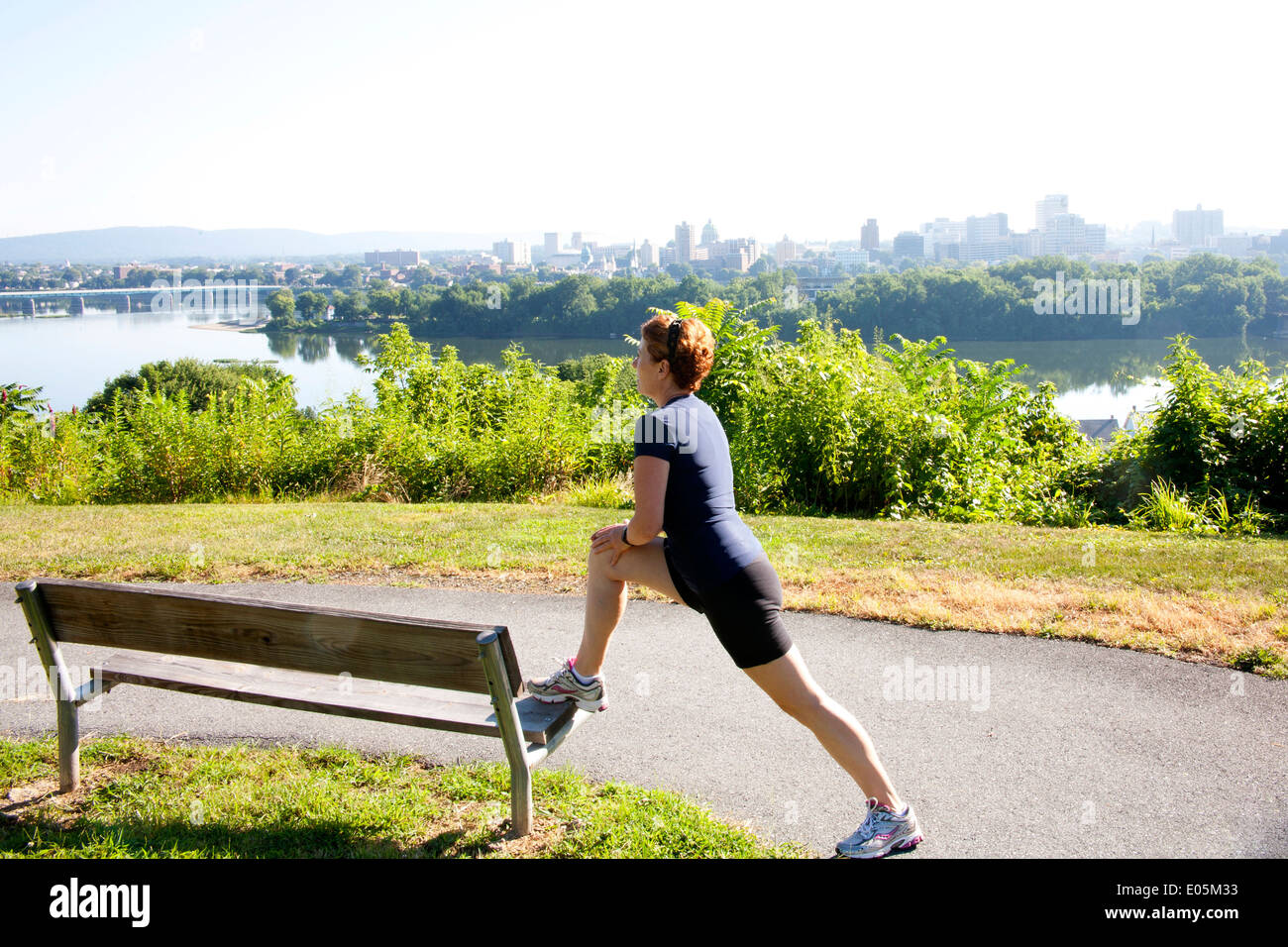 Female jogger stretching on a bench before running along a path overlooking  a riverview city skyline on a hazy, hot summer day Stock Photo - Alamy