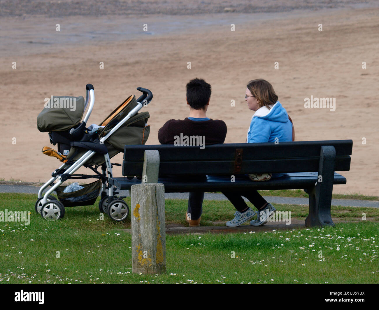 Young couple sat on a public bench with a pram, Bude, Cornwall, UK Stock Photo