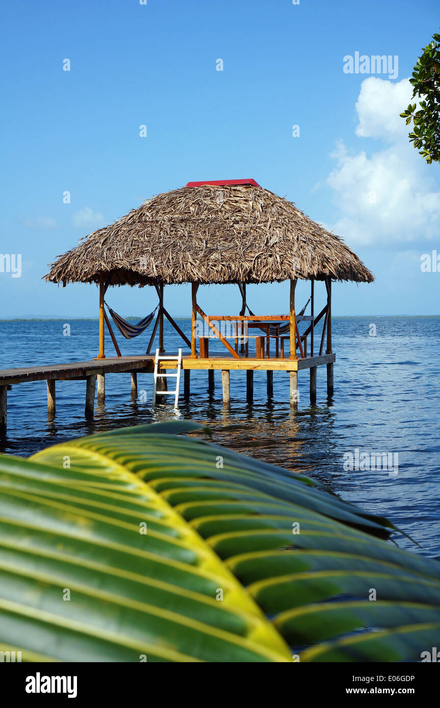 Tropical hut over water with thatched roof and a palm leaf in foreground Stock Photo