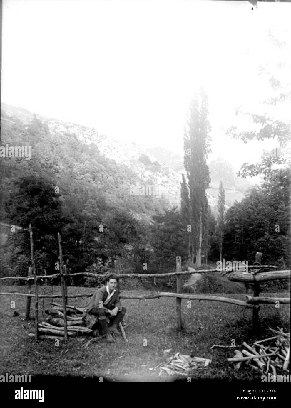 Homme accroupi devant une barrière en bois en constructrion et un tas de bois coupé Stock Photo