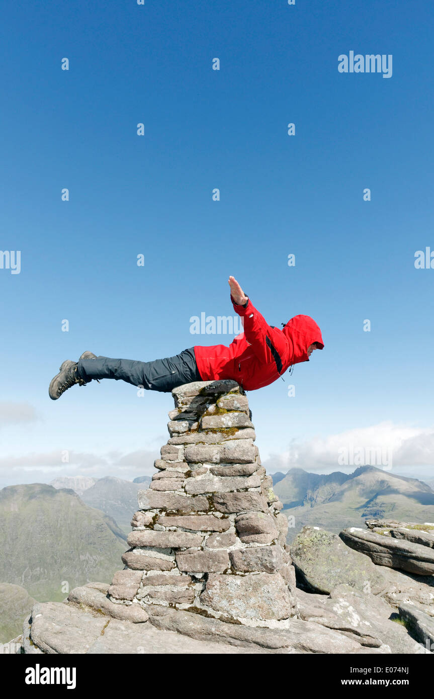 A hillwalker in a red jacket pretending to be superman, flying on the summit cairn of Beinn Alligin in Torridon, Scotland Stock Photo