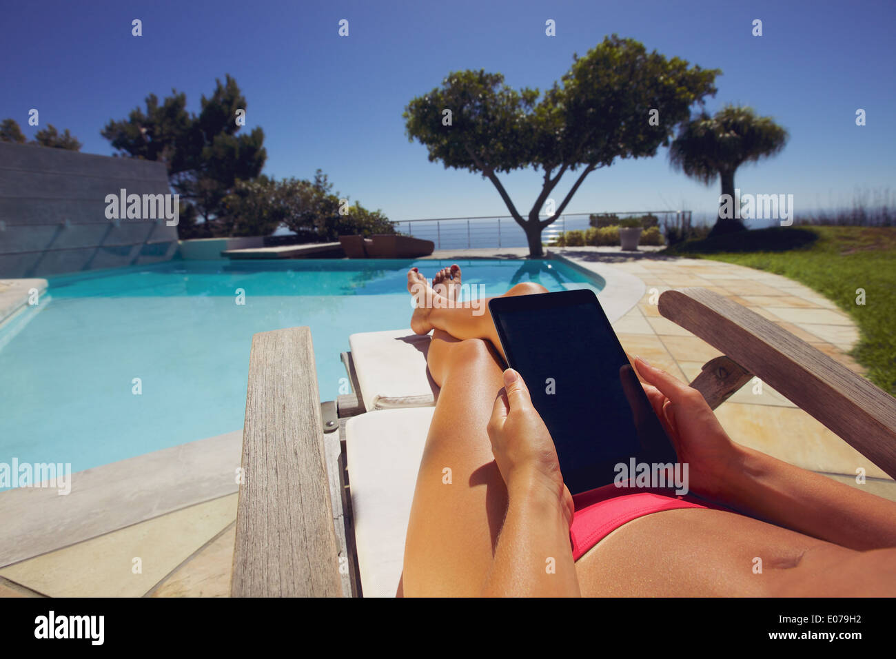 Relaxed woman using digital tablet by the swimming pool. Tanned female model sunbathing and holding tablet PC at poolside. Stock Photo