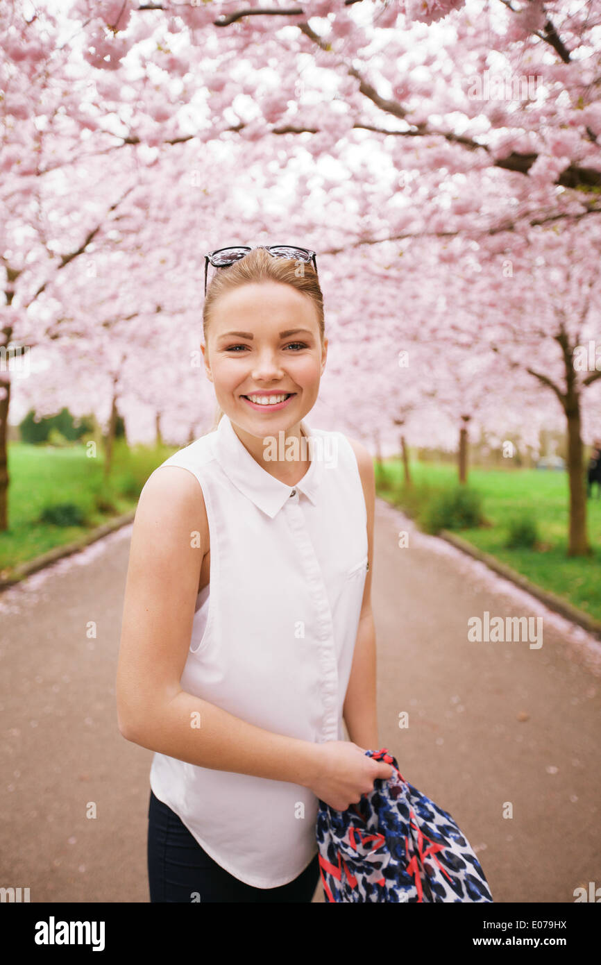 Happy young woman looking at camera while at spring blossom park. Female model on pathway through cherry blossom trees. Stock Photo