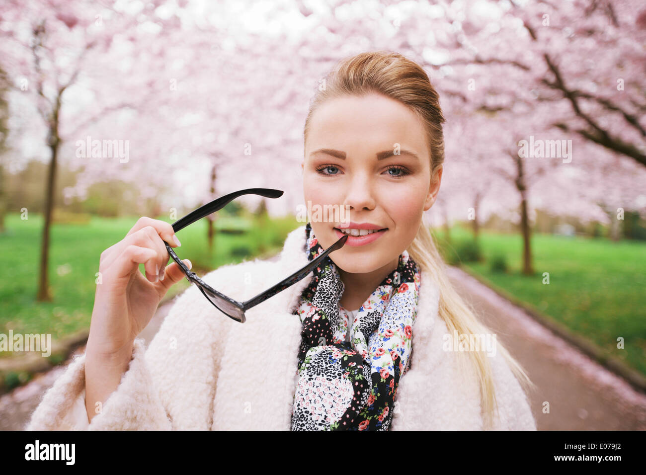 Pretty young woman at spring park holding sunglasses looking at camera. Caucasian female model posing at spring blossom park. Stock Photo