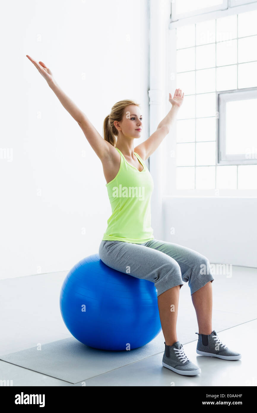 Young woman sitting on exercise ball, arms raised Stock Photo