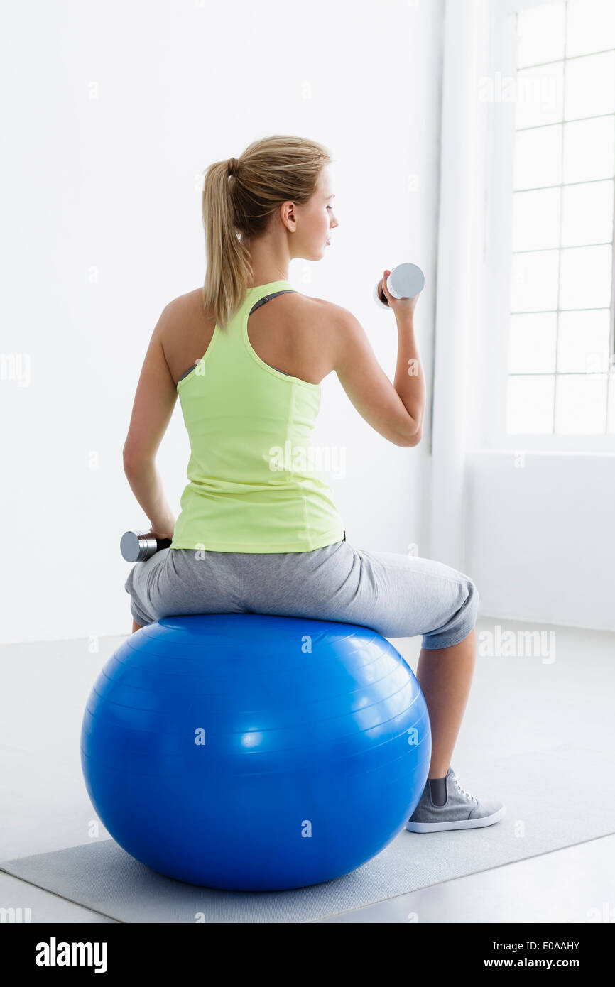 Young woman sitting on exercise ball, rear view, lifting weights Stock