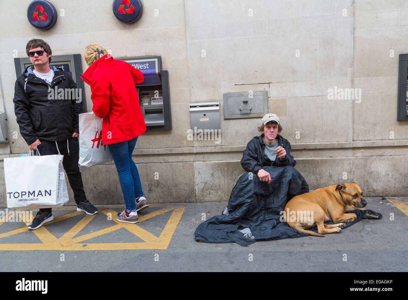 Young millennials withdraw cash from an ATM whilst a homeless man and his dog sit nearby begging for money, London England UK Stock Photo