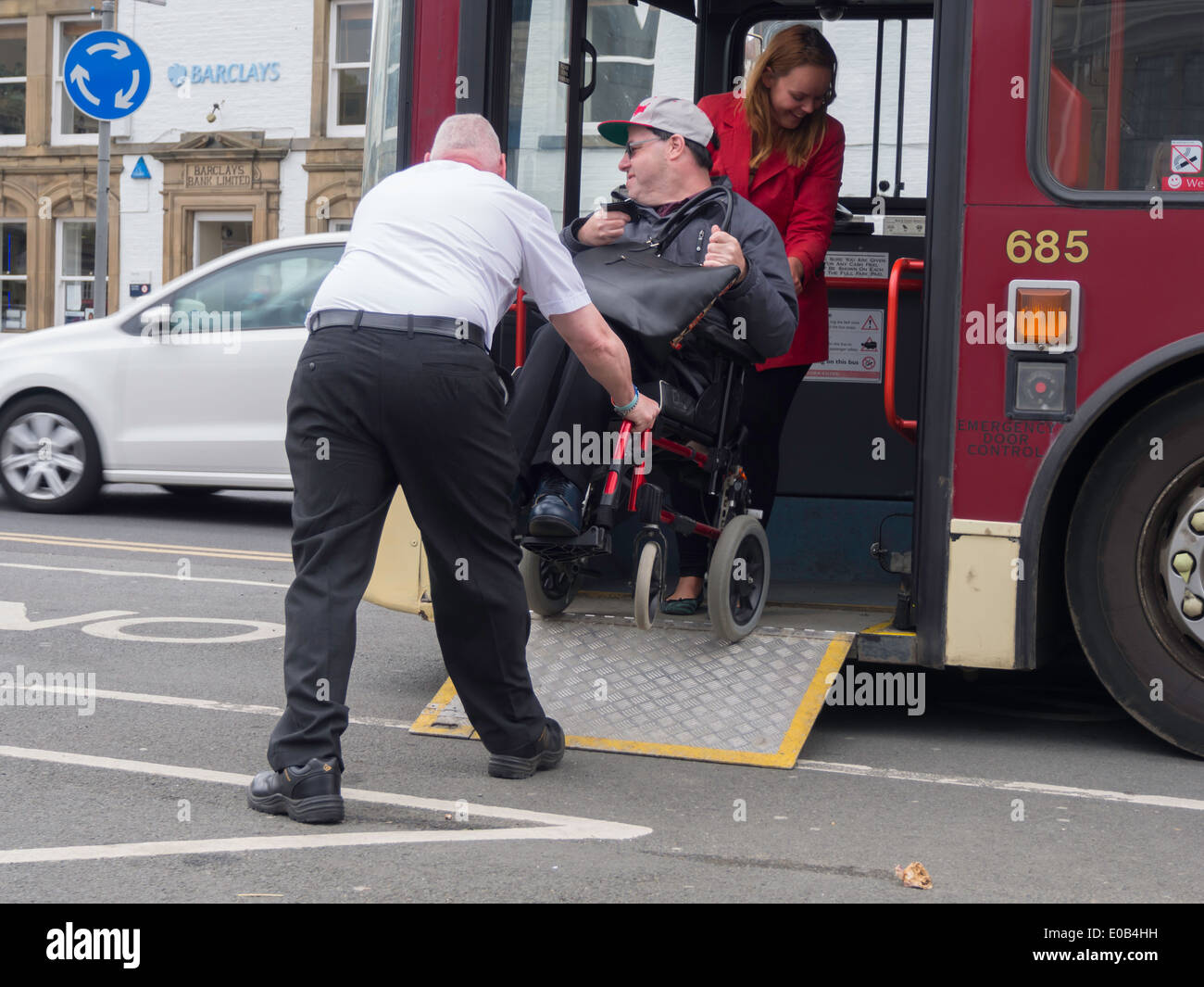 A disabled man in a wheelchair with his carer being helped to board a bus by its driver Stock Photo
