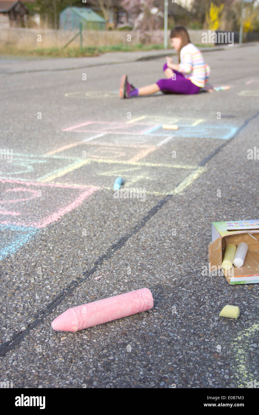 Hopscotch drawing with coloured crayon on asphalt, girl sitting in the background Stock Photo