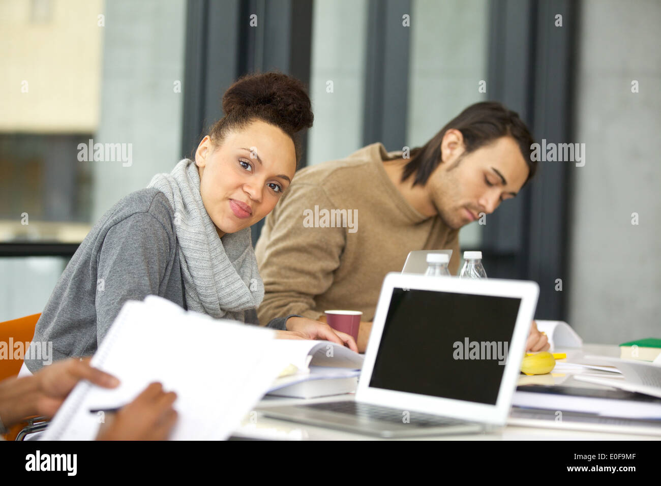 Confident young woman sitting at table looking at camera. Students studying with books and laptop in library. Stock Photo
