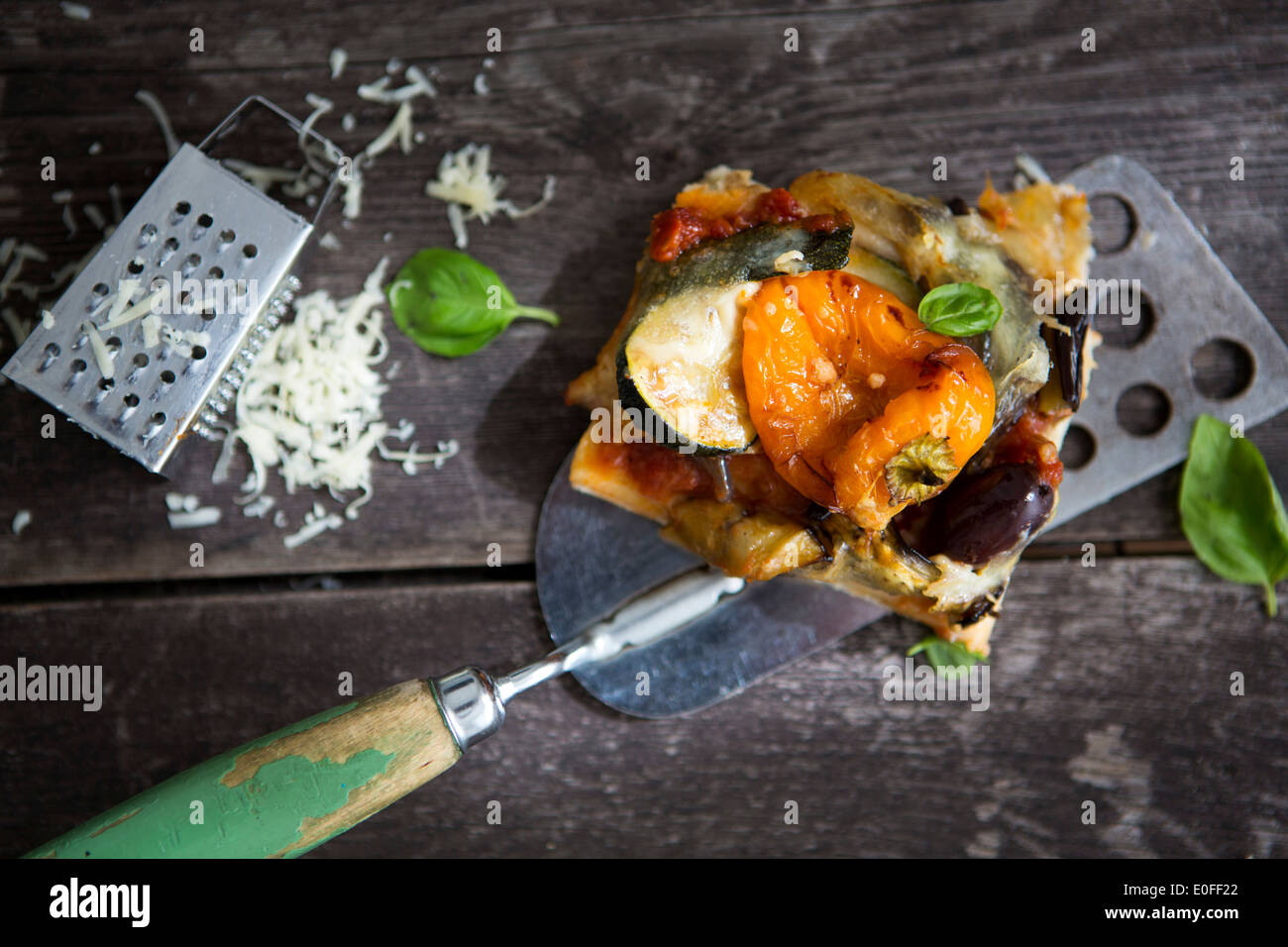 A slice of homemade pizza with pepper, courgette, olive and basil and a cheese grater Stock Photo