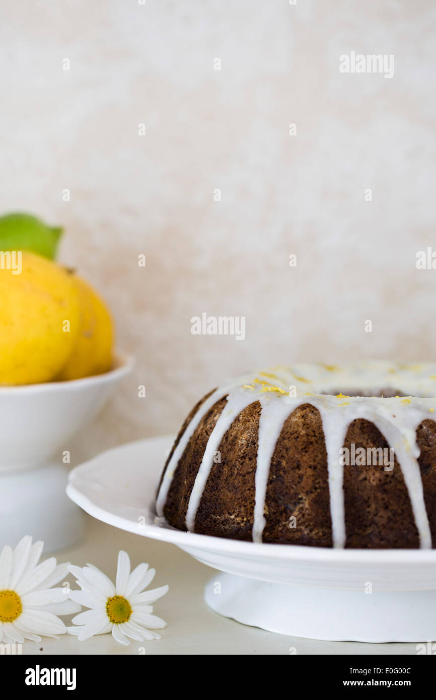 Round mould iced date and walnut cake served on white plate, with lemons in background Stock Photo