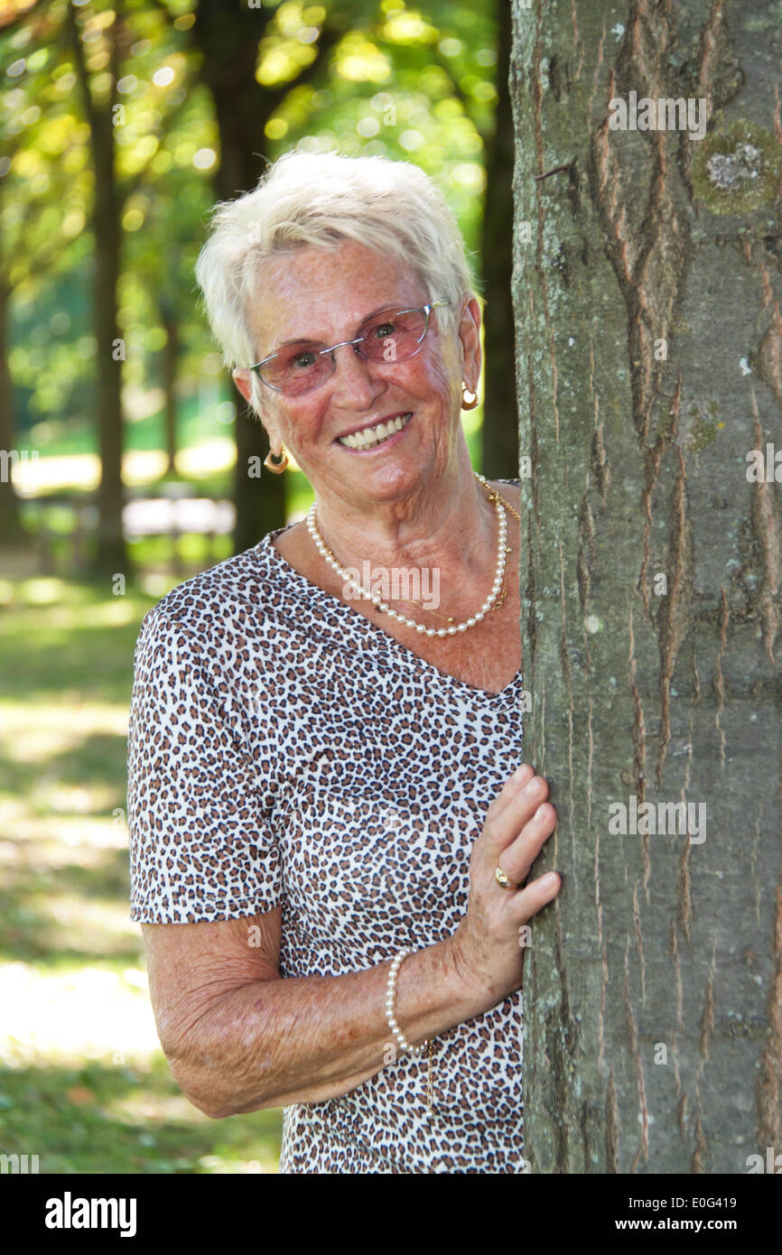 Happy older senior stands in a tree, Froehliche aeltere Seniorin steht an einem Baum Stock Photo