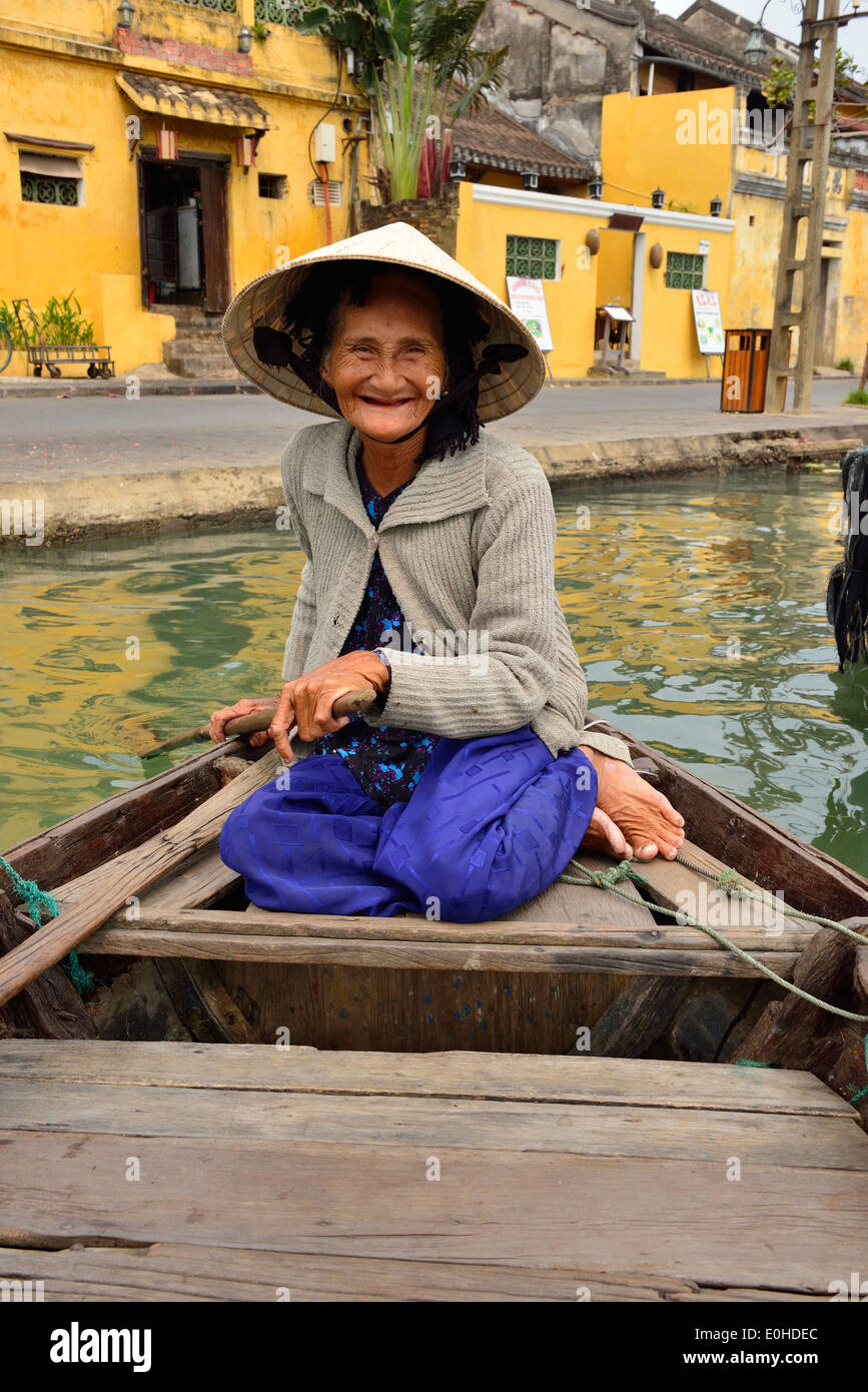 Elderly smiling Vietnamese woman taking tourists for rides on the river in the town of Hoi An, Vietnam, paddling from the stern of her river boat. Stock Photo