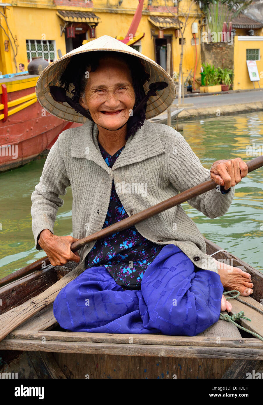 Elderly smiling Vietnamese woman taking tourists for  rides on the river in the town of Hoi An, Vietnam paddling from the stern of her river boat. Stock Photo