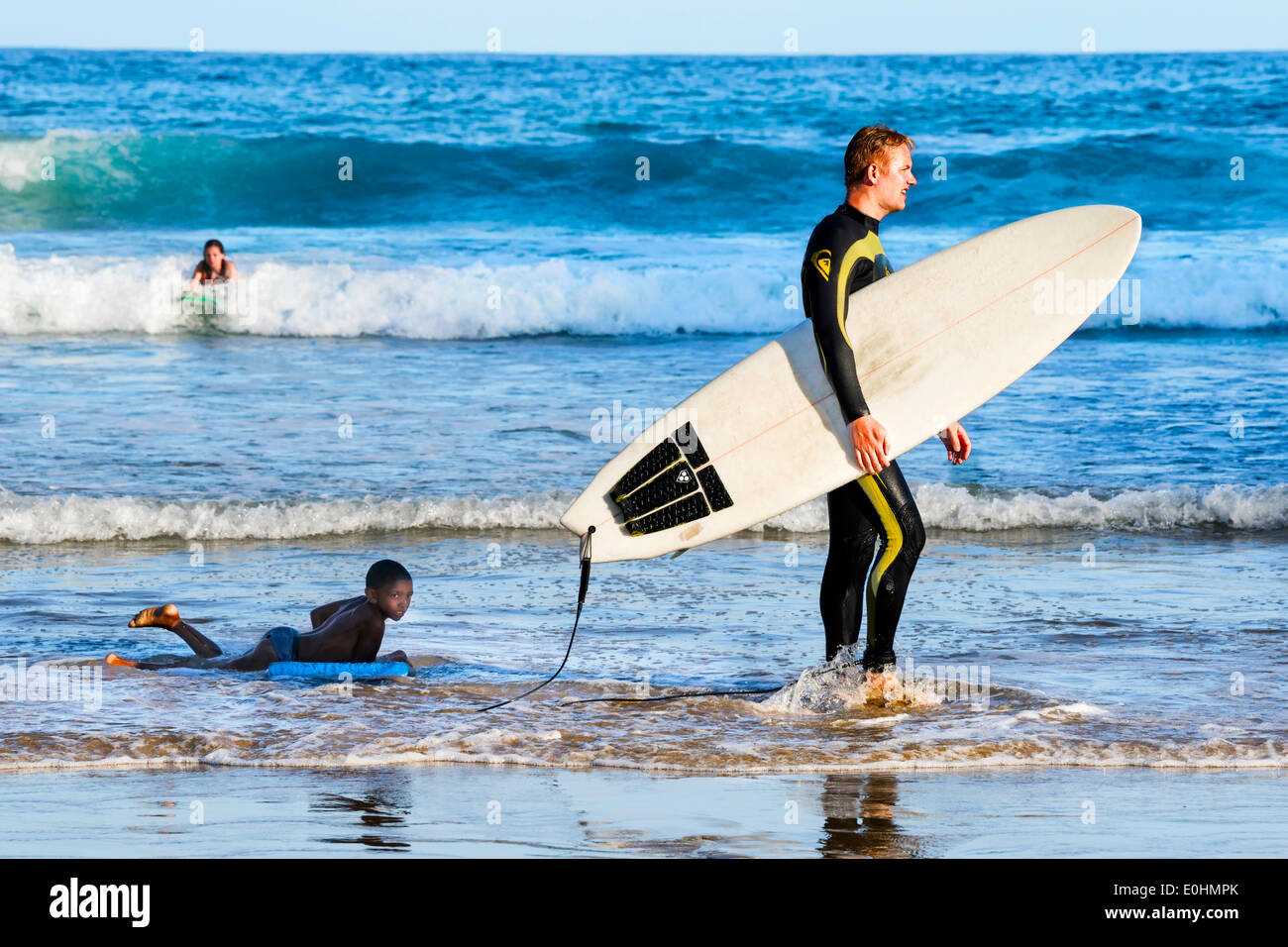 Surfing beach scene of man holding surfboard, boy on body board behind