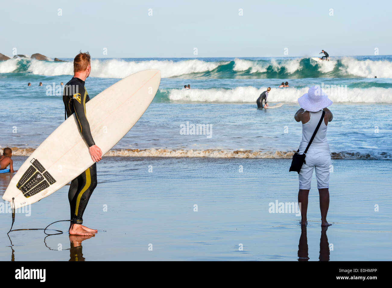 Beach scene with man holding surfboard and woman watching surfers